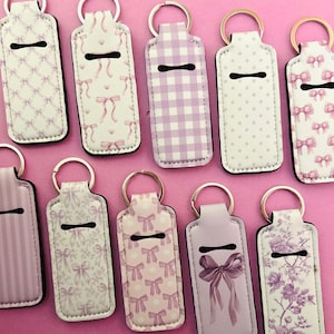 May include: A selection of keychains featuring bow, polka dot, gingham, and floral patterns. The keychains are in pink, white, and purple, with a black trim and a silver key ring. The designs are varied.
