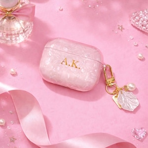 May include: A pink and white pearlescent AirPod case with gold-coloured hardware and a charm. The case has the initials "A.K." in gold. The background is pink with decorative elements like pearls and a ribbon.