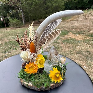 May include: A woodland-themed centerpiece featuring a deer antler, dried flowers, moss, and crystals on a wooden base.