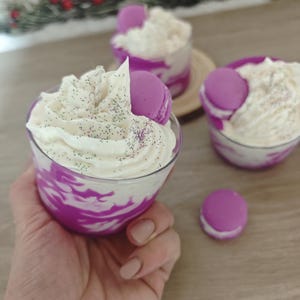 Puede incluir: Tres copas de postre con crema batida blanca, remolinos morados y un macaron morado. Las copas están sobre una superficie de madera, con una ramita de vegetación en el fondo. La crema batida está espolvoreada con purpurina verde y morada.