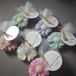 Puede incluir: Una colección de recuerdos en forma de margarita en varios colores pastel, cada uno envuelto en una red blanca y atado con hilo. Cada recuerdo tiene una etiqueta circular con el texto "Georgia & Augustin 31.08.2025" y un diseño de arcoíris.