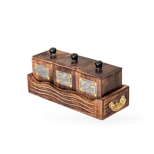 Puede incluir: Un juego de tres recipientes rectangulares de madera con acabado de veta de madera marrón oscuro, cada uno con una placa de metal plateado etiquetada como Sugar, Coffee o Tea. Los recipientes tienen pomos negros y se asientan en una bandeja de madera a juego con asas doradas.