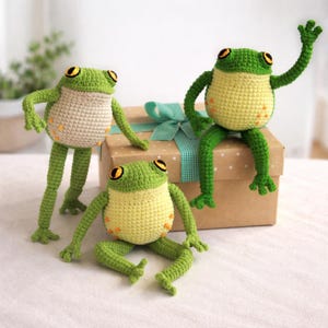 May include: Three handmade crochet frog toys in various poses. The frogs have green heads and limbs, yellow bellies, and tan bodies. They have large, round eyes with black pupils and orange accents. One frog sits on a brown gift box with a teal ribbon.