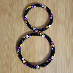 Puede incluir: Dos pulseras de cuentas negras con un patrón de cuentas moradas, amarillas y blancas. Las pulseras son circulares y están sobre una superficie de madera.