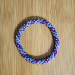 May include: A beaded bracelet in a circular shape. The bracelet is made of small, colorful beads in shades of purple and light blue. The beads are arranged in a pattern, creating a textured surface. The bracelet is displayed on a wooden surface.