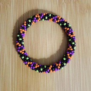 May include: A beaded bracelet in a circular shape, featuring a pattern of black, orange, purple, and green beads. The bracelet is set against a wooden background, showcasing the vibrant colors of the beads.