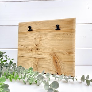 Könnte beinhalten: Ein rechteckiges, hellbraunes Holzbrett mit einer ausgeprägten natürlichen Holzmaserung und zwei kleinen schwarzen Metallklammern an der Oberkante zum Befestigen von Gegenständen. Die Holzoberfläche zeigt sichtbare Astlöcher und Texturen.