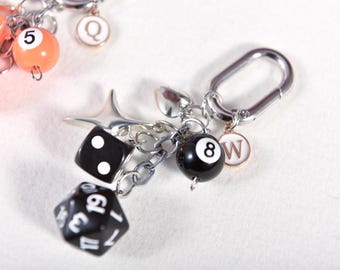 May include: A silver keychain with various charms, including a black eight ball, a black die with white dots, and a black and white twenty-sided die. Additional charms include a star, a letter "W", and a number "5" on an orange ball.