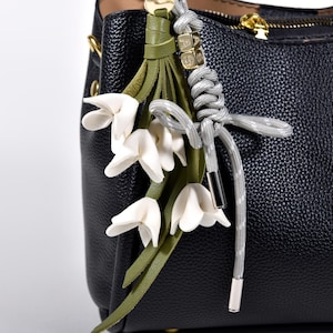 May include: A black leather handbag with a decorative floral charm. The charm features white flower blossoms, green leaves, and a gray cord with silver-toned metal accents. The bag has a gold zipper.