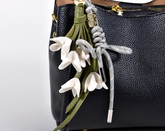 May include: A black leather handbag with a decorative floral charm. The charm features white flower blossoms, green leaves, and a gray cord with silver-toned metal accents. The bag has a gold zipper.