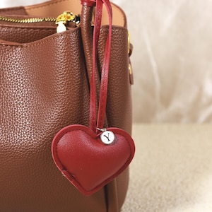 Puede incluir: Un bolso de mano de cuero marrón con un charm en forma de corazón rojo. El charm tiene una inicial "Y" plateada y está sujeto al bolso con una correa de cuero roja. El bolso tiene cierre de cremallera y herrajes dorados.