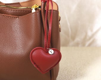 May include: A brown leather handbag with a red heart-shaped charm. The charm has a silver-colored initial "Y" and is attached to the bag with a red leather strap. The bag has a zipper closure and gold-colored hardware.
