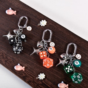 Może przedstawiać: Trzy srebrne breloczki do kluczy z różnymi zawieszkami, w tym kostkami do gry, kulami bilardowymi i zawieszkami w kształcie gwiazd. Kostki są czarne, pomarańczowe i zielone. Tło to ciemna drewniana powierzchnia z małymi elementami dekoracyjnymi.