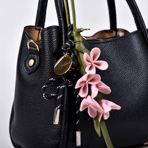 Könnte beinhalten: Schwarze Lederhandtasche mit strukturierter Oberfläche. Die Tasche hat einen dekorativen floralen Akzent mit rosa Blüten und einem goldfarbenen Anhänger. Die Griffe sind stabil und die Tasche hat ein klassisches, strukturiertes Design. Ein stilvolles Accessoire für den täglichen Gebrauch.