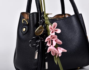 May include: Black leather handbag with a textured finish. The bag features a decorative floral accent with pink flowers and a gold-toned charm. The handles are sturdy, and the bag has a classic, structured design. A stylish accessory for everyday use.