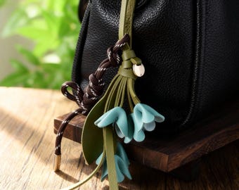 May include: A leather bag charm featuring a cluster of blue flower accents and a green leather leaf design. The charm includes a brown and white rope detail and a gold-colored metal accent.
