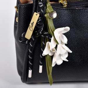 Könnte beinhalten: Schwarze Lederhandtasche mit strukturierter Oberfläche. Die Tasche hat einen goldenen Reißverschluss und einen dekorativen Anhänger mit dem Wort "Custom". Ein Cluster aus weißen Blumen und grünen Blättern ist am Griff befestigt.
