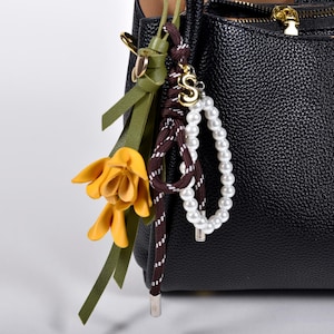 Könnte beinhalten: Schwarze Lederhandtasche mit strukturierter Oberfläche. Ein dekorativer Anhänger hängt am Griff, mit einer gelben Blume, grünen Bändern, einem braun-weißen Seil und einem Perlenakzent mit dem goldenen Buchstaben "S".