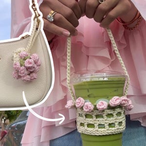 May include: A cream-colored crochet cup sleeve featuring a lattice pattern and a row of small pink crocheted flowers along the top edge, attached to a matching crochet carrying strap, holding a clear plastic cup filled with a green iced beverage.