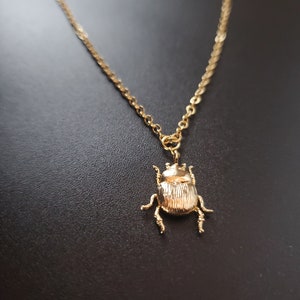 May include: A gold-coloured necklace with a detailed scarab beetle pendant. The beetle hangs from a delicate chain, creating an elegant accessory. The necklace is displayed against a dark background.