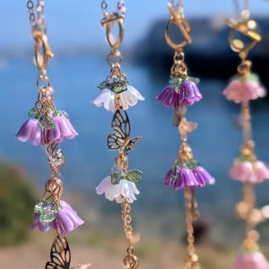 May include: Four gold-toned necklaces featuring dangling bell-shaped flower charms in shades of purple and white, accented with small green leaves and delicate gold-toned butterfly cutouts. Each necklace includes a gold-toned toggle clasp closure.