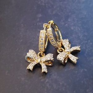 May include: A pair of gold-colored hoop earrings with bow-shaped pendants. The hoops and bows are encrusted with small, clear crystals, creating a sparkling effect. The earrings are displayed against a dark, textured background.