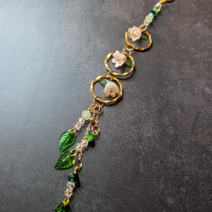 May include: A gold-toned jewelry piece featuring three circular accents with white flower details and green leaves. The design includes dangling green leaf charms and clear and green beads, creating an elegant, nature-inspired accessory.