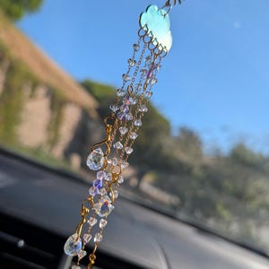 May include: A car charm featuring a sunburst and cloud design. The charm is made of gold-colored metal and features clear, iridescent crystal beads and teardrop-shaped pendants. The cloud is a light blue color. The charm hangs from a gold chain.