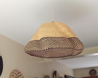 Pode incluir: Uma luminária pendente tecida com uma parte superior bege natural e uma parte inferior marrom estampada pende do teto. Cestas de parede tecidas decorativas em vários tamanhos e designs adornam a parede. Uma mesa de jantar com um vaso tecido e flores está em primeiro plano.