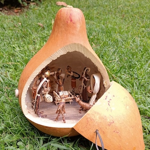 Può includere: Un presepe artigianale racchiuso all'interno di una zucca svuotata. La zucca è di colore marrone chiaro, con un'apertura a cerniera che rivela un diorama in miniatura di figure. Le figure sono realizzate con materiali naturali e raffigurano la scena della natività biblica.