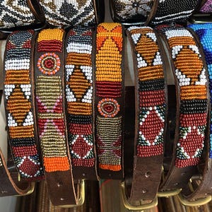 May include: A collection of beaded dog collars with brown leather straps. The collars feature intricate patterns in various colors, including red, orange, white, blue, and black. The collars are displayed with their buckles visible.