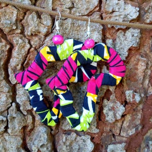 May include: A pair of dangling earrings featuring irregular, multi-sided shapes wrapped in fabric with a vibrant geometric pattern in hot pink, lime green, black, and yellow. Each earring is accented with a single bright pink bead at the top.