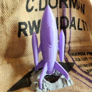 Könnte beinhalten: Ein lilafarbenes Plastik-Raketenspielzeug mit einem grauen Sockel. Die Rakete hat eine spitze Nase und zwei seitliche Flügel. Der Sockel ist strukturiert, um wie eine Mondlandungsstelle auszusehen.