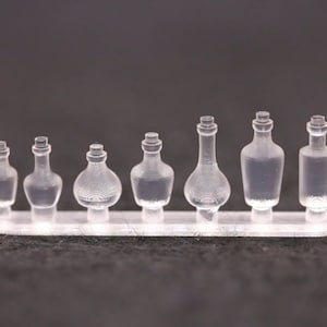 Könnte beinhalten: Ein Set aus sieben durchsichtigen Glasflaschen in verschiedenen Formen und Größen. Die Flaschen sind in einer Reihe auf einem durchsichtigen Kunststoffsockel angeordnet.