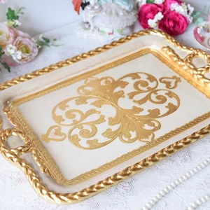 Può includere: Vassoio decorativo rettangolare color crema con un design a filigrana dorata. Il vassoio ha un bordo e manici dorati a forma di corda. Un design a filigrana dorata è centrato sulla superficie del vassoio. Il vassoio viene probabilmente utilizzato per servire o esporre.