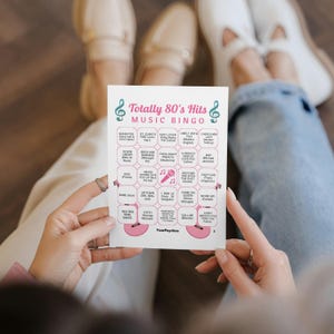 Pu&ograve; includere: Una scheda di gioco rettangolare bianca intitolata Totally 80's Hits Music Bingo con note musicali color ottanio, un'icona di microfono rosa e illustrazioni di chitarre rosa. La griglia contiene venticinque riquadri con vari titoli di canzoni.