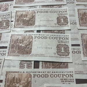 Peut inclure: Un gros plan sur une pile de coupons alimentaires vintage du d&eacute;partement de l'agriculture des &Eacute;tats-Unis. Les coupons sont blancs avec du texte noir et pr&eacute;sentent une image de la D&eacute;claration d'ind&eacute;pendance. Chaque coupon vaut un dollar.