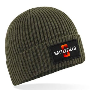 Könnte beinhalten: Olivgrüne Strickmütze mit Rippenstruktur. Ein schwarzes Rechteck zeigt das Wort "BATTLEFIELD" in Weiß, mit einem stilisierten orangefarbenen "O" in der Mitte. Ein warmer, lässiger Kopfschmuck.