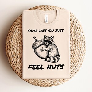May include: A beige t-shirt with a black illustration of a raccoon holding a nut. The text "Some days you just feel nuts" is printed on the shirt.