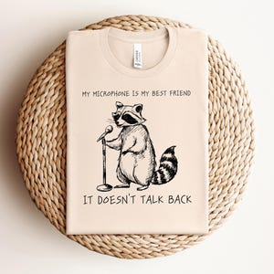 May include: A beige t-shirt with a black and white illustration of a raccoon holding a microphone. The text "My microphone is my best friend. It doesn't talk back." is printed on the shirt.