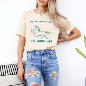 May include: A light beige t-shirt featuring a graphic of a unicorn running with a clock in its hoof. The text on the shirt reads "The only exercise I get is running late."