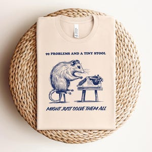 May include: A beige t-shirt with a blue illustration of a mouse sitting on a stool and typing on a typewriter. The text "99 Problems and a Tiny Stool Might Just Solve Them All" is printed on the shirt.