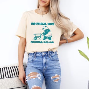 Puede incluir: Una camiseta beige claro con un gráfico de un panda empujando una carretilla. El texto "Another Day Another Dollar" está impreso en la camiseta.