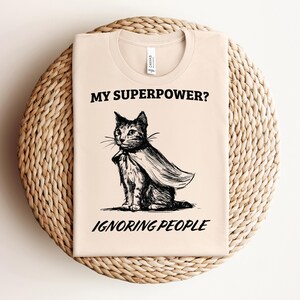 May include: A beige t-shirt with a black and white illustration of a cat wearing a cape. The text "MY SUPERPOWER? IGNORING PEOPLE" is printed on the shirt.