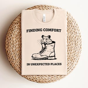 May include: A beige t-shirt with a black graphic of a hamster wearing a boot. The text "Finding Comfort in Unexpected Places" is printed below the graphic.