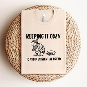 May include: A beige t-shirt with black text that reads "Keeping it cozy to avoid existential dread". A cartoon turtle wearing a hat and scarf is pulling a sled.