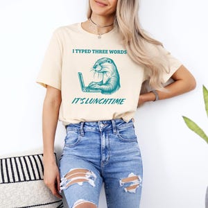May include: A light beige t-shirt with a teal graphic of an otter using a laptop. The text on the shirt reads "I typed three words it's lunchtime".