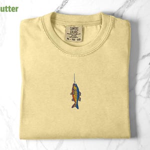 May include: A yellow t-shirt with a small embroidered image of a fish on a fishing line. The shirt has a tag that says "Comfort Colors" and "XL - TG - EG".