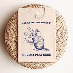 May include: A beige t-shirt with a blue illustration of a possum wearing a jester's hat and juggling apples. The text "Not sure if I should dance or just play dead" is printed on the shirt.