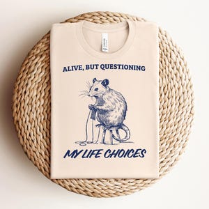 May include: A beige t-shirt with a blue graphic of a rat sitting on a stool and holding a spool of thread. The text "Alive, but questioning my life choices" is printed in blue below the graphic.